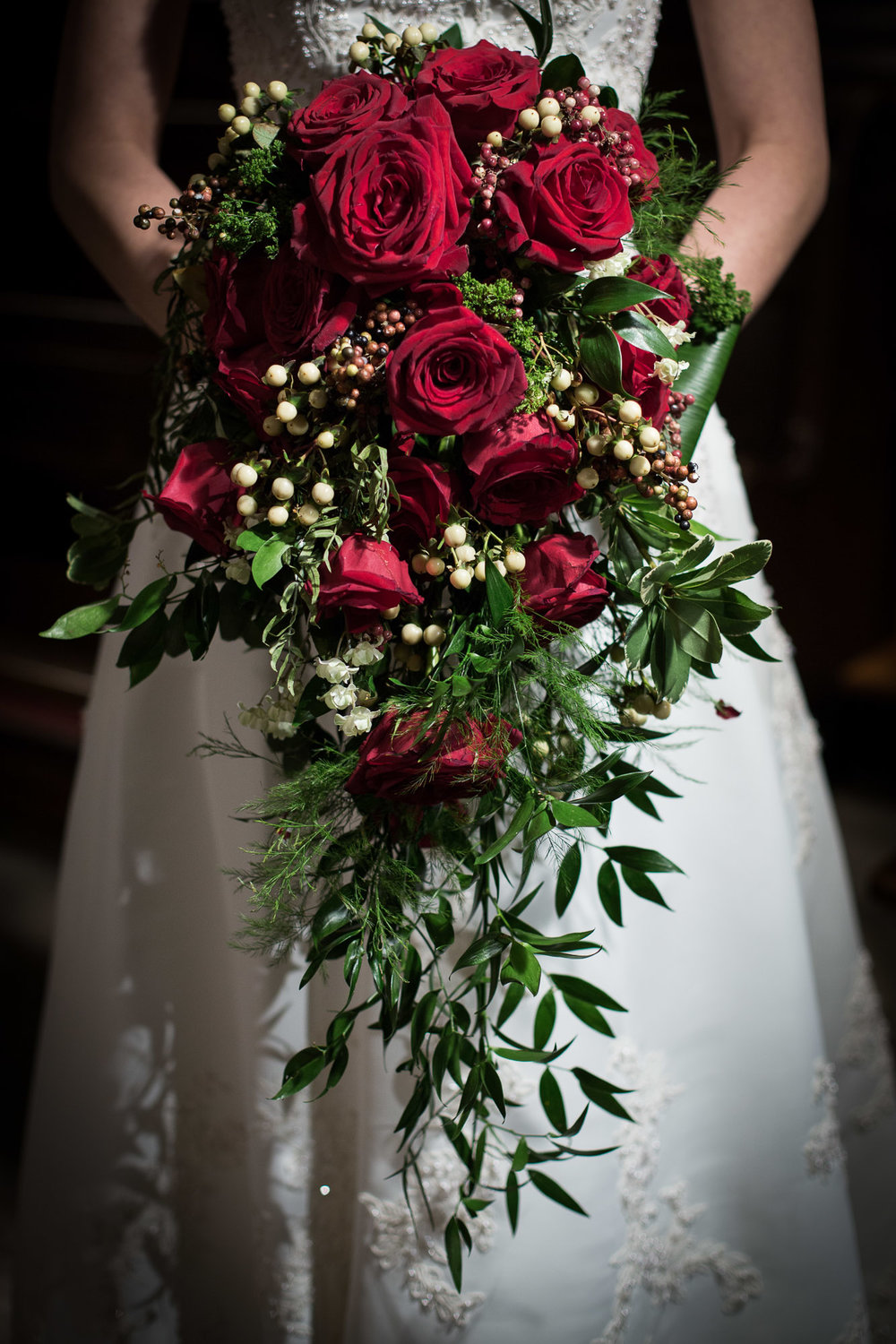 Cascading bouquet of red roses, white berries. The greenery is a mix of Israeli ruscus leaves and soft Asparagus Ferns